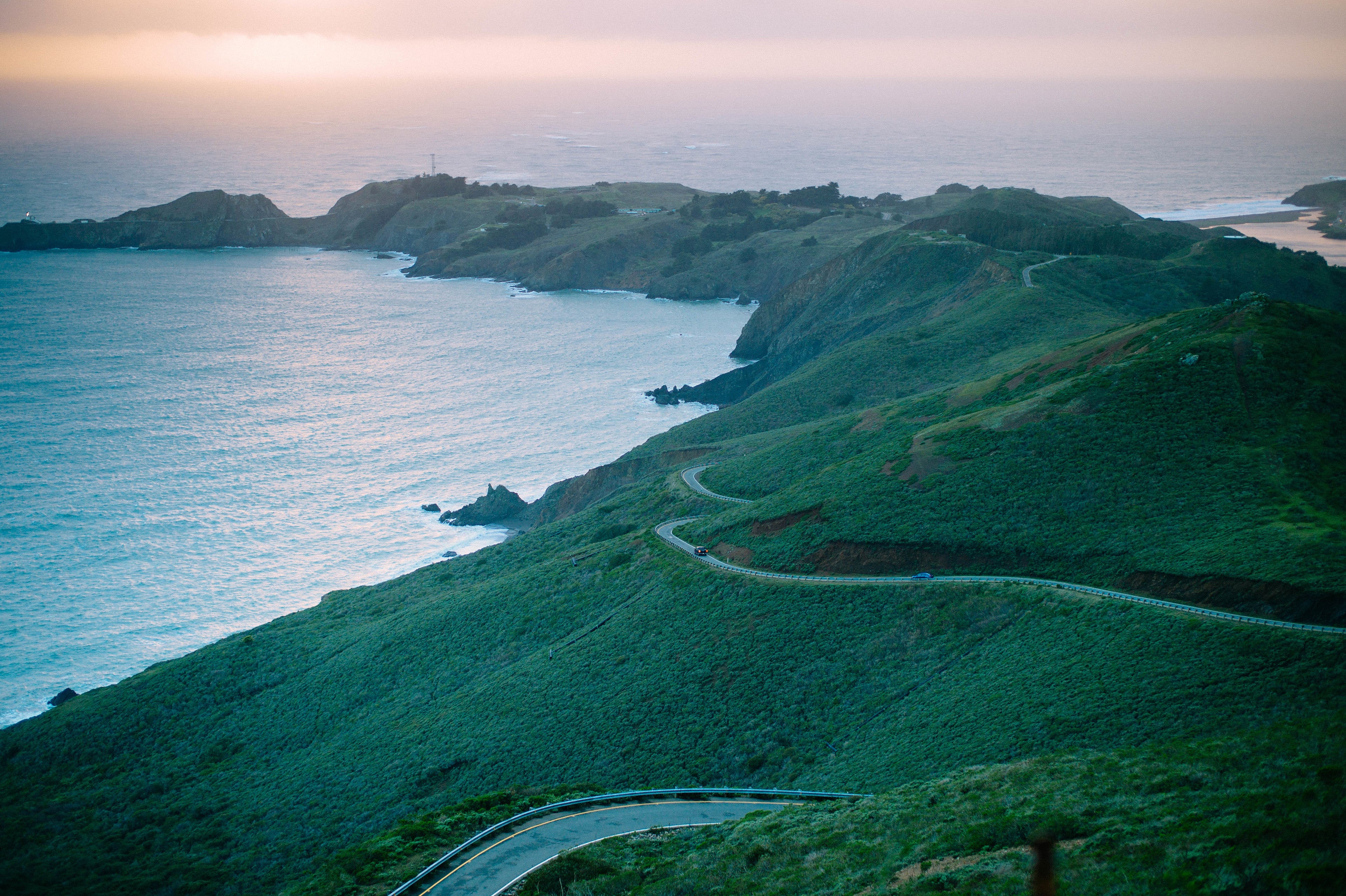 an aerial photo of a highway winding througn the hills, next to the coast, with a sunset in the background. there is a car travelling away from the camera in the distance